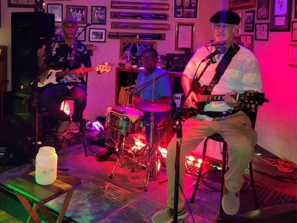A live musical performance featuring three musicians plays in a cozy, decorated venue in Roatan, Honduras, with framed photographs and memorabilia on the wall.