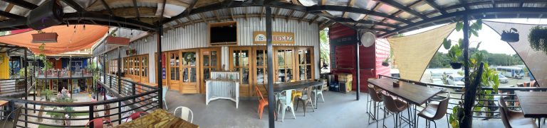 A panoramic view of a rooftop bar and dining area in Roatan, Honduras, featuring wooden tables, colorful chairs, hanging plants, shades, and a view of parked cars and greenery outside.