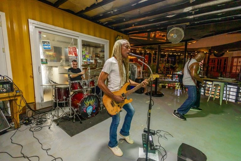 A live musical performance with a guitarist, drummer, and vocalist performing indoors at a restaurant or bar in Roatan, Honduras, with colorful signage and a vibrant atmosphere.