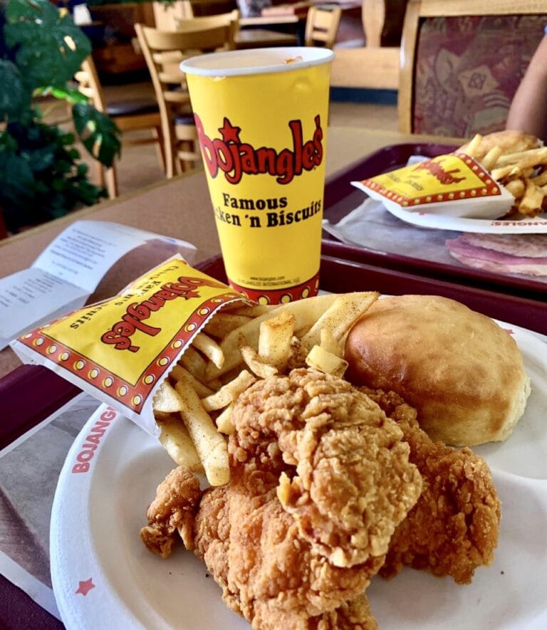 A plate of fried chicken, French fries, and a dinner roll is shown with a D'Angelo's cup and a tray of more chicken and fries in the background, set on a table in a casual dining restaurant on Roatan, Honduras.