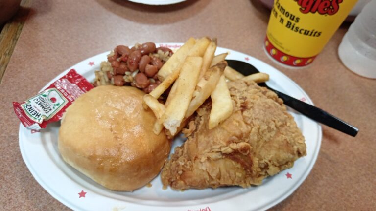 A plate of typical Southern comfort food including fried chicken, French fries, baked beans, and a dinner roll, with a Roatan, Honduras-themed soda and condiments on a countertop.