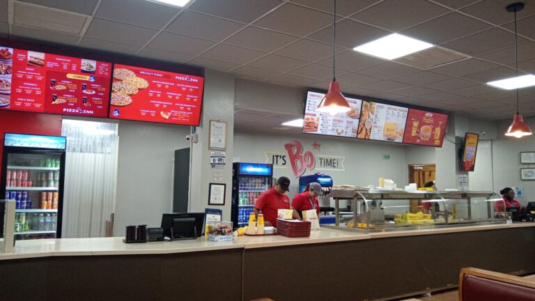 Inside a fast-food restaurant on Roatan, Honduras, employees serve customers behind a counter with digital menu boards displaying pizza, combos, and beverages, while a refrigerator stocked with soda cans is visible to the left.