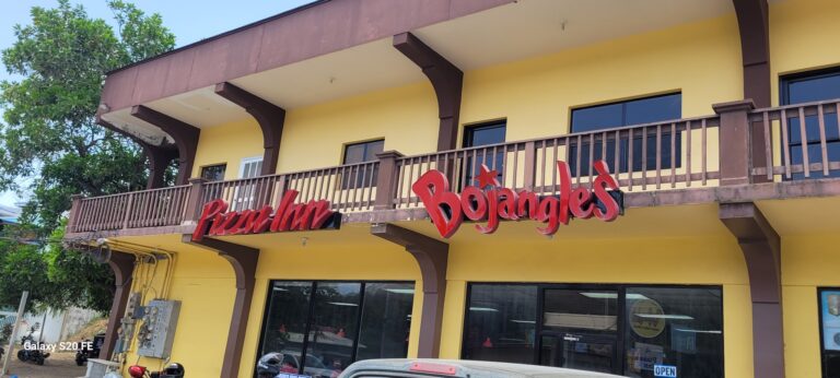 The yellow two-story building in Roatan, Honduras, features a wooden balcony with a red "Pura Vida Bungalows" sign and large glass windows on the ground floor, with lush green trees visible in the background.
