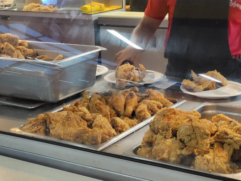 Fried chicken pieces displayed in a glass case at a food establishment on Roatan, Honduras, with a worker preparing plates behind the counter.