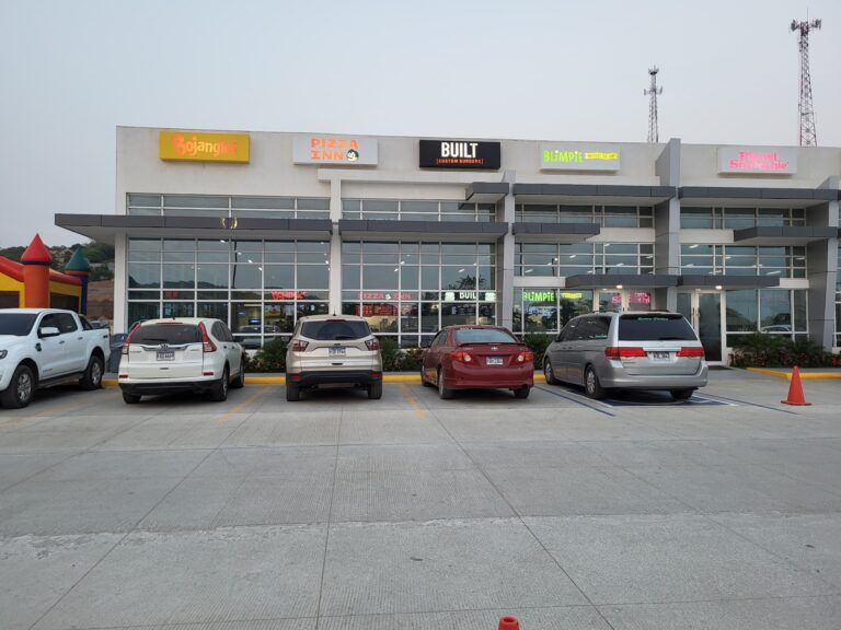 A multi-level commercial building in Roatan, Honduras features signage for various eateries including Bojangles, Pizza Inn, Built Custom Burgers, Blimpie, and Planet Smoothie, with a parking lot and cars in the foreground.
