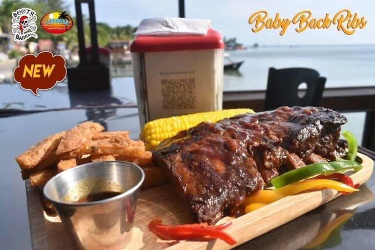A plate of baby back ribs with barbecue sauce, corn on the cob, fried plantains, and sliced colorful peppers is served on a wooden tray at a seaside restaurant in Roatan, Honduras, with a waterfront view in the background.