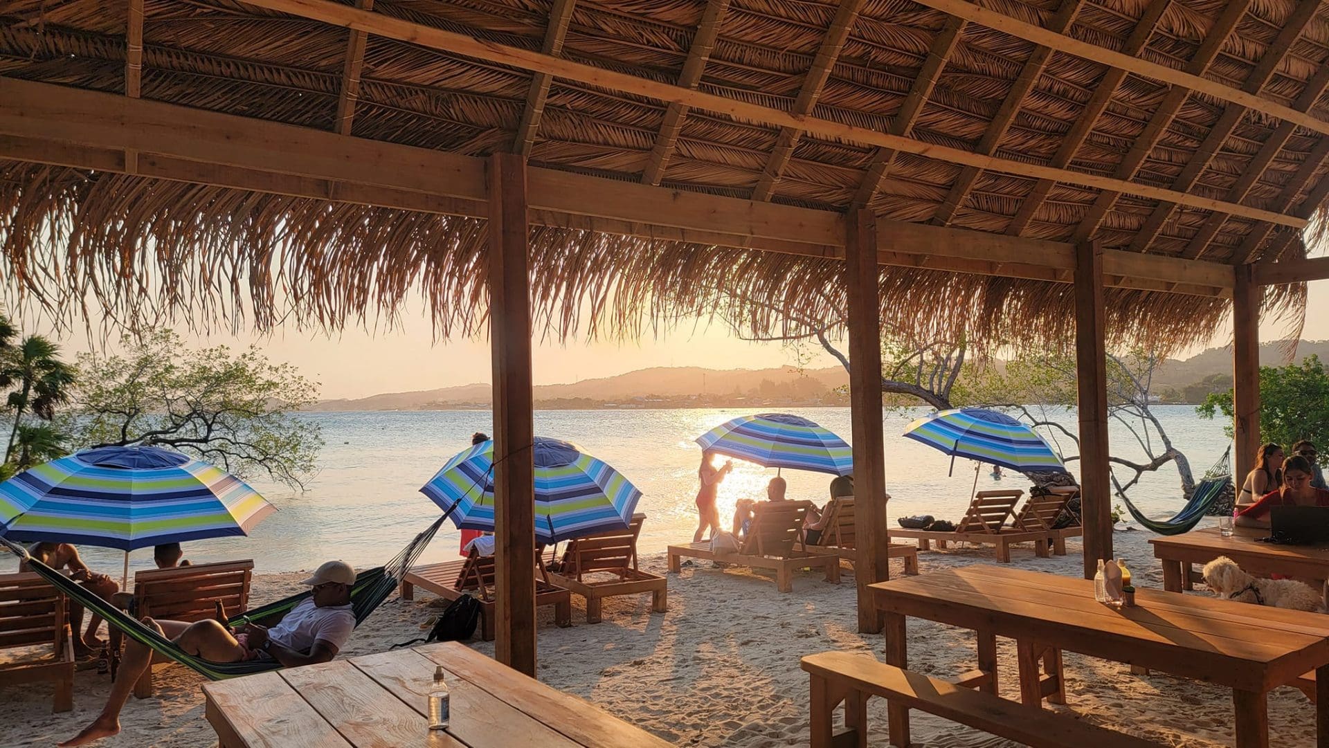 People relax under a thatched-roof structure on the sandy beach of Roatan, Honduras, with colorful umbrellas, lounge chairs, hammocks, and the ocean visible during sunset.