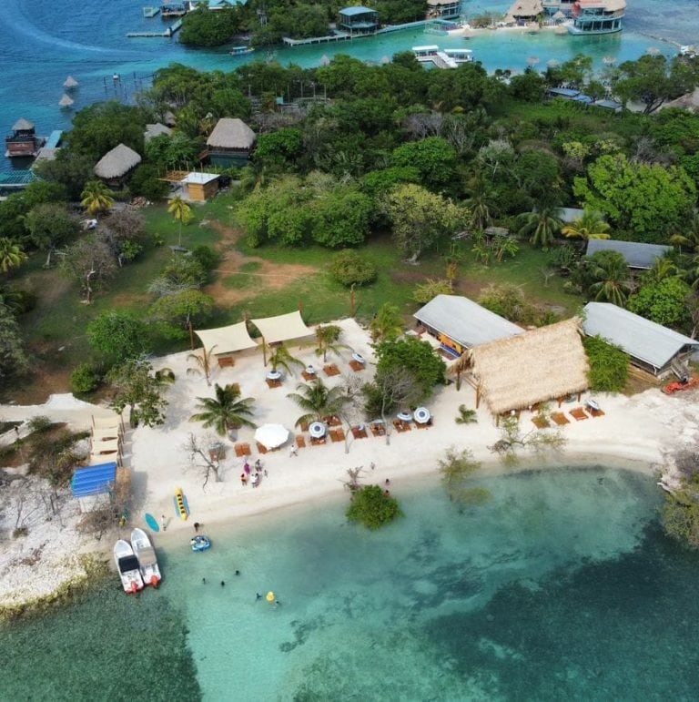 Aerial view of a tropical beach resort in Roatan, Honduras, featuring a sandy shoreline with lounge chairs, shaded cabanas, palm trees, clear turquoise water for swimming, and lush green vegetation surrounding several thatched-roof and open-air structures near the water.