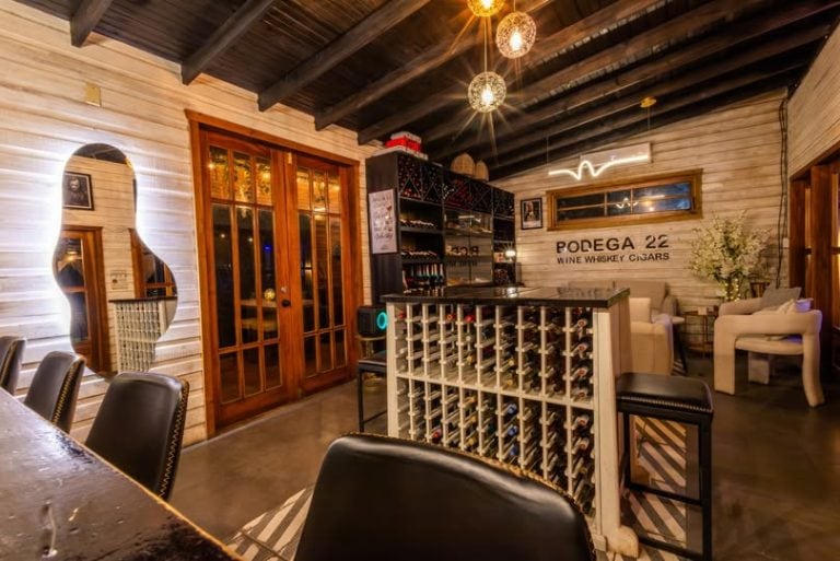 A cozy interior of a wine and cigar lounge on Roatan in Honduras features a wooden ceiling, a wine display, seating with white armchairs and black leather chairs, decorative lighting, and a sign for "Podega 22 Wine Whiskey Cigars" on the wall.