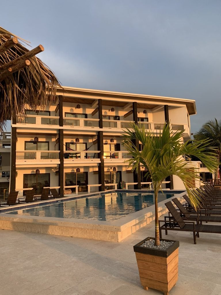 A modern hotel building with multiple balconies overlooks a swimming pool surrounded by lounge chairs and potted palm trees on Roatan, Honduras.