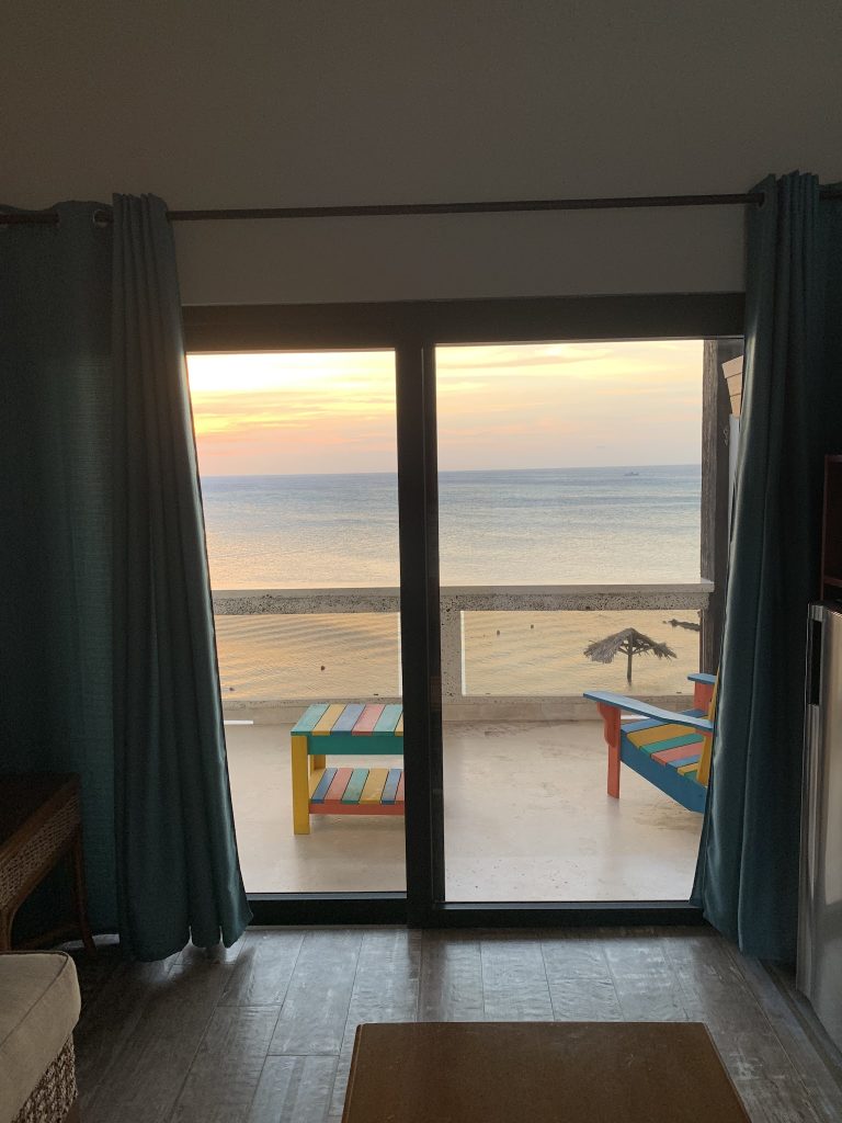 A view from inside a room in Roatan, Honduras, looking out through glass sliding doors at a balcony with colorful furniture, overlooking the ocean during sunset.