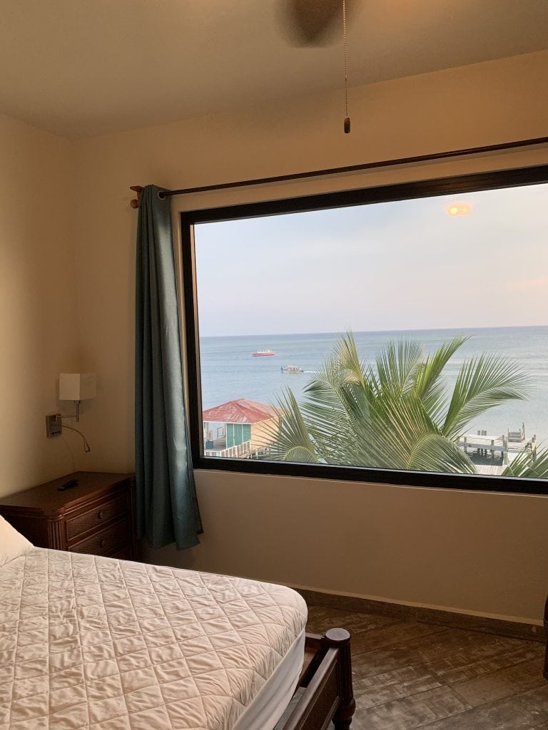 A bedroom with a large window overlooking the ocean and palm trees in Roatan, Honduras, featuring a wooden bedside table, a lamp, and a bed with a quilted beige cover.