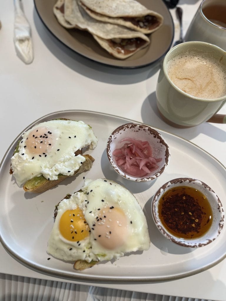 A breakfast plate with eggs benedict topped with black sesame seeds, served with small bowls of pickled red onions, salsa, and hot sauce, alongside a plate of crêpes filled with fruit or chocolate, on a white tray in Roatan, Honduras.