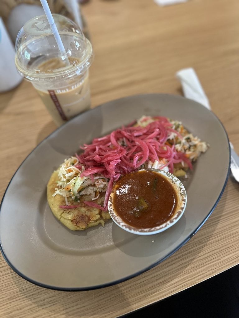 A plate of Honduras-style food with shredded cabbage, pickled pink onions, a small cup of spicy red sauce, and cornmeal cake, with an iced coffee in a plastic cup with a straw on a wooden table in Roatan, Honduras.