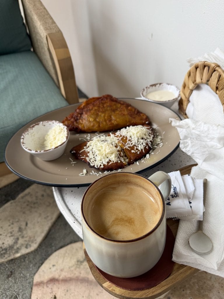 A cup of coffee, a plate with a slice of tamale topped with cheese, a small bowl of sour cream, and a basket of tissues are arranged on a small round table in a casual setting on Roatan, Honduras.