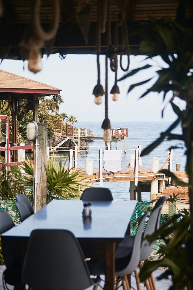 A dining area with black chairs and a table overlooking a wooden deck and the ocean in Roatan, Honduras, featuring tropical plants and hanging light fixtures under a thatched roof.