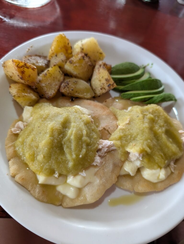 A plate of Honduras-style comfort food featuring baked chicken topped with cheese sauce, fried plantains, and fresh sliced green plantains, set on a white dish on a wooden table in Roatan, Honduras.