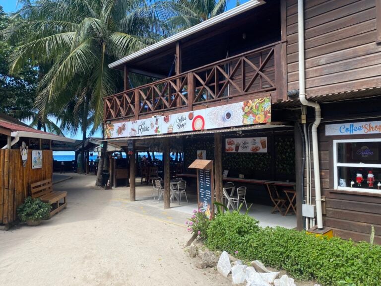 A beachfront coffee shop in Roatan, Honduras features a wooden building with outdoor seating under a shaded area, surrounded by palm trees and tropical plants.