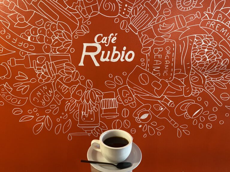 A white coffee cup filled with black coffee rests on a saucer with a spoon on a red wall decorated with white line drawings of coffee-related items at Café Rubio in Roatan, Honduras.