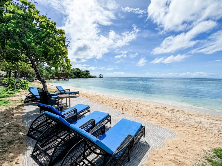 A row of blue lounge chairs facing the Caribbean Sea along a sandy beach with green trees, wooden pier structures, and a partly cloudy sky over Roatan in Honduras.