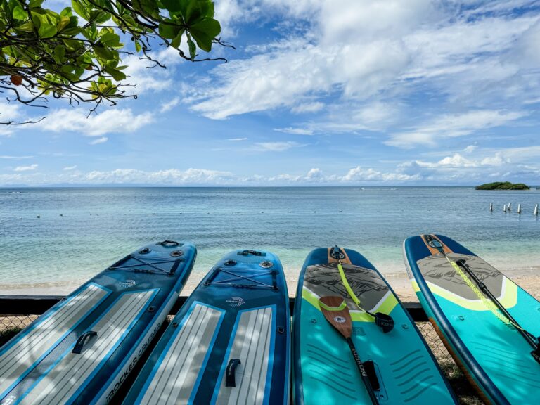 Stand-up paddleboards are lined up on the beach in Roatan, Honduras, with the ocean and a partly cloudy sky in the background.