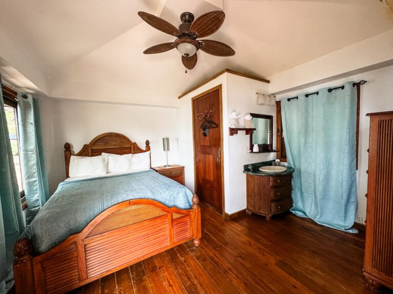 A bedroom in a Roatan, Honduras hotel features a wooden bed with a blue patterned bedspread, a wooden dresser with a turquoise curtain covering a window, and a ceiling fan with palm leaf-shaped blades.
