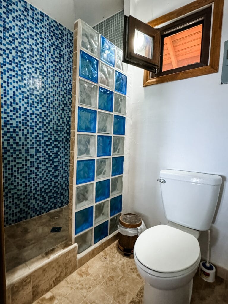 A small bathroom featuring a tiled shower area with blue mosaic tiles, a partition wall made of glass blocks in shades of blue, and a wooden-framed window providing natural light, located in Roatan, Honduras.