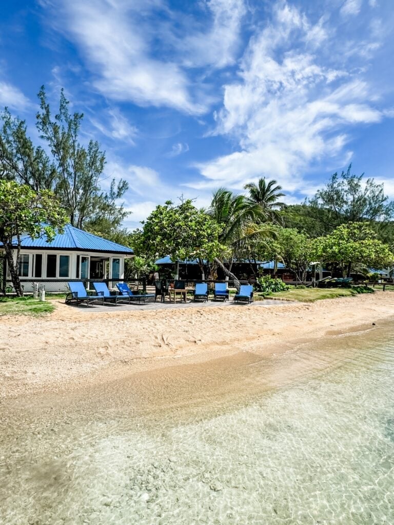A sandy beach in Roatan, Honduras, features lounge chairs, a small building with a blue roof, lush green trees, and a clear sky with scattered clouds.