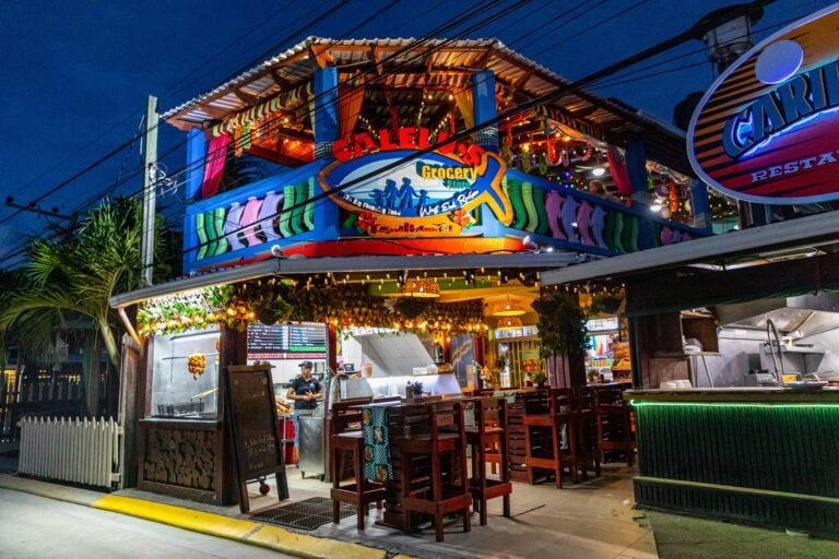 A brightly lit, colorful seafood restaurant and grocery store in Roatan, Honduras, features an outdoor seating area with wooden tables, a neon sign, and vibrant decorations under a night sky.