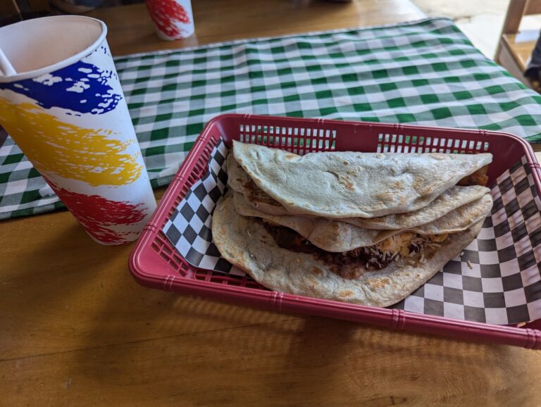 A basket of folded quesadillas on a checkered paper liner, a paper cup with colorful stripes, and a green and white checkered tablecloth on a wooden table in Roatan, Honduras.