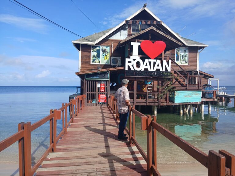 A man walks along a wooden pier leading to a two-story waterfront building in Roatan, Honduras, featuring a large sign that says "I ♥ Roatan" and sports-themed artwork on the exterior walls.