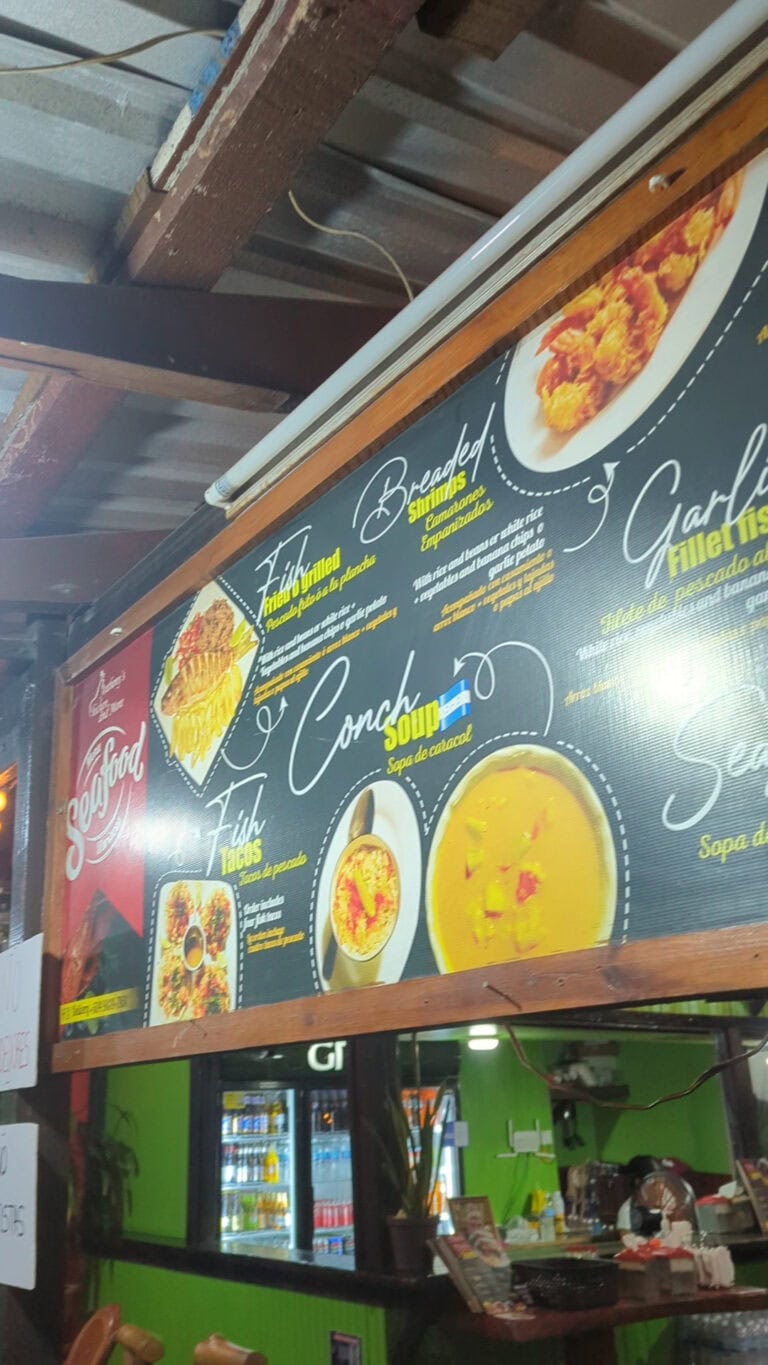 A restaurant menu board displaying fish, shrimp, chicken, and soup dishes is mounted on a wooden frame inside a Roatan, Honduras eatery, with a green counter and a refrigerator filled with beverages visible in the background.