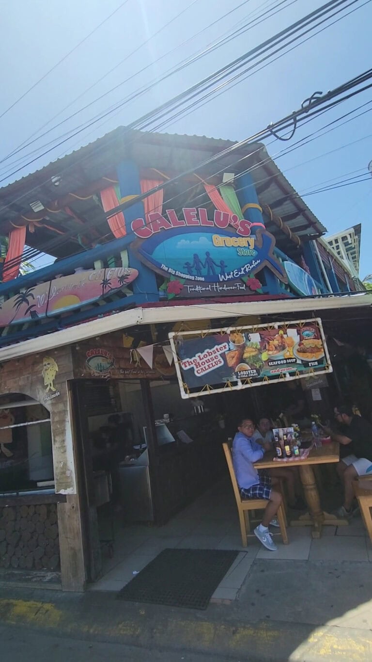 A two-story restaurant in Roatan, Honduras named Calelu's Greasy Store features colorful signage and outdoor seating with people dining at a wooden table on the sidewalk.