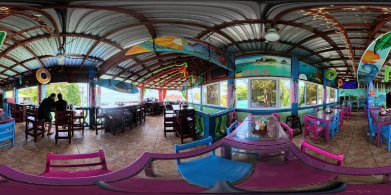 A colorful tropical-themed restaurant interior in Roatan, Honduras, featuring vibrant pink and blue chairs, large windows with views of palm trees and the ocean, and ocean-inspired decorations including murals and hanging surfboard artwork.