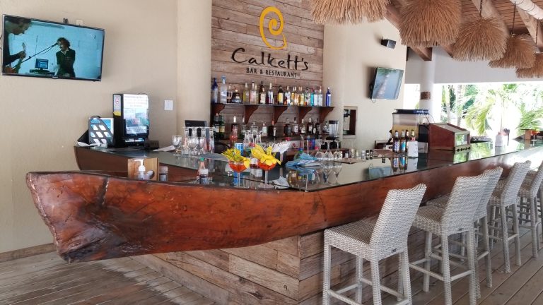 A bar and restaurant in Roatan, Honduras, features a wooden boat-shaped bar with glassware, bottles, and tropical cocktails, with wicker chairs, a wooden wall with the "Calkett's Bar & Restaurant" sign, and tropical decor including thatched roof features.