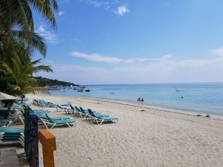 A sandy beach in Roatan, Honduras, featuring rows of blue and white lounge chairs under palm trees, with boats anchored in calm turquoise waters under a clear blue sky.