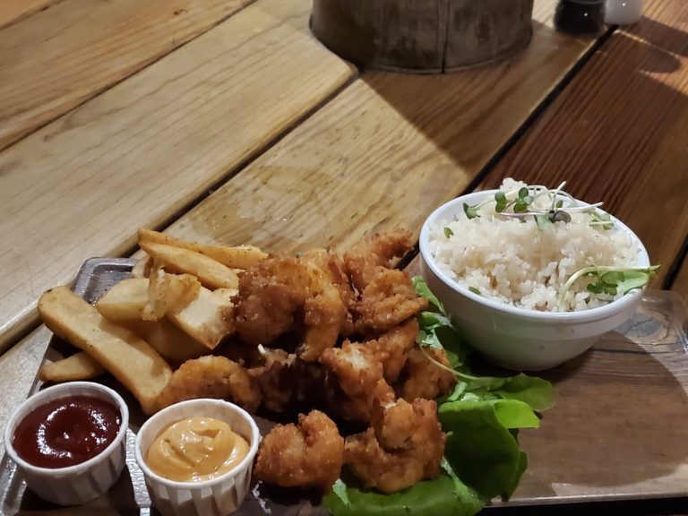 A plate of fried fish, French fries, and toasted bread is served with small bowls of ketchup, mustard, and dipping sauces on a wooden table at a restaurant in Roatan, Honduras.