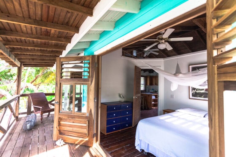 An interior view of a wooden bungalow in Roatan, Honduras, featuring a bedroom with a white bed, a blue dresser, wooden shutters, ceiling fans, and a balcony with lush tropical greenery outside.