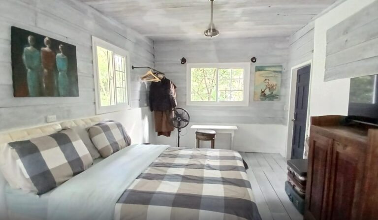 A cozy bedroom on Roatan, Bay Islands, Honduras featuring whitewashed wooden walls, a large bed with checkered bedding, and a small window overlooking lush greenery outside.