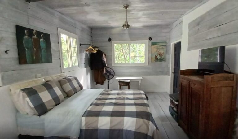 A cozy bedroom in Roatan, Honduras, featuring light-colored wooden walls, a large bed with checked bedding, and a small window overlooking lush greenery outside.