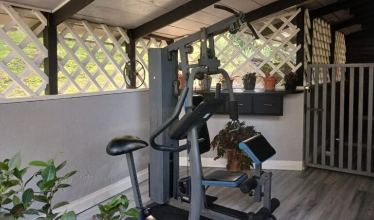Indoor gym area in Roatan, Bay Islands, Honduras featuring exercise equipment with a wooden ceiling and lattice window view of lush greenery outside.