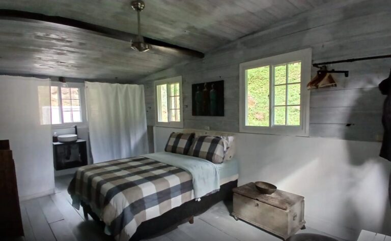 Interior view of a cozy bedroom in a rustic cottage on Roatan, Honduras, featuring a wooden bed with checkered bedding, small windows with green scenery outside, and simple decor including a painting and a wooden chest.