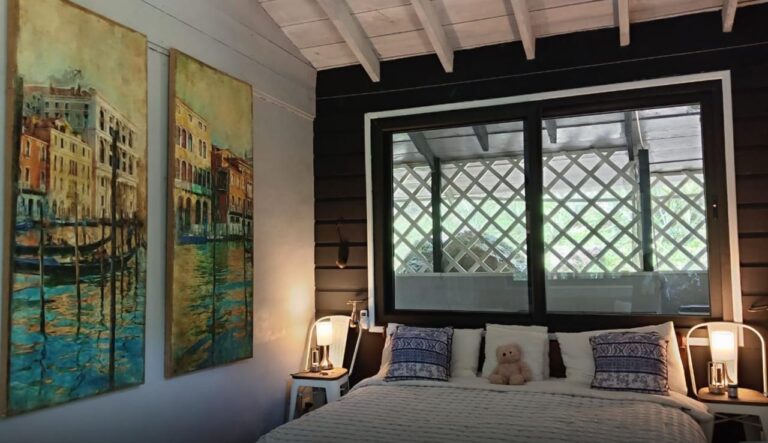 A cozy bedroom in a Roatan accommodation features a large bed with white linens, blue patterned pillows, and a stuffed teddy bear. The room has a black wooden accent wall behind the bed, illuminated by warm bedside lamps, and a large window with a view of a balcony with a lattice railing and greenery outside.