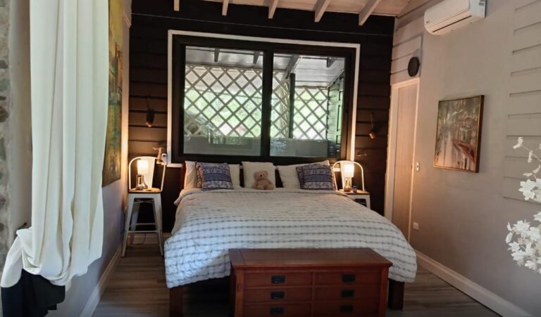 A cozy bedroom in Roatan, Honduras, featuring a large bed with decorative pillows, soft lighting from bedside lamps, and a window with a view of lush greenery outside.