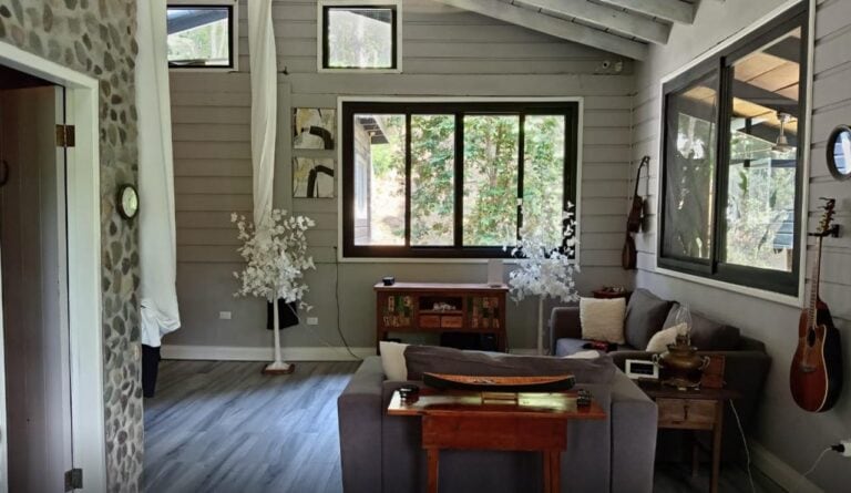 Interior view of a cozy living room in a Roatan, Honduras home featuring white wooden walls, large windows with natural light, and comfortable gray sofas decorated with throw pillows.