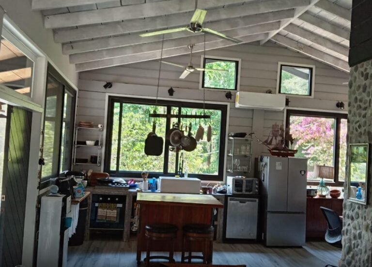 Inside a modern kitchen with white wooden beams ceiling, large windows showing lush greenery and pink flowering trees from Roatan, Honduras, and a hanging pot rack with cookware.