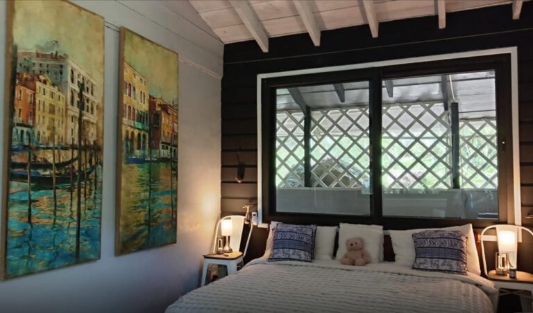 A cozy bedroom in a Roatan, Honduras beachfront cottage with nautical themed artwork on the wall and a large window overlooking a balcony with a lattice railing.