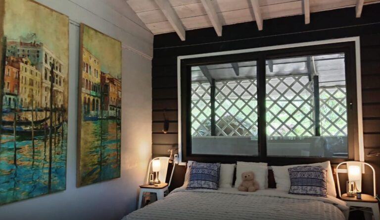 A cozy bedroom in Roatan, Honduras, featuring a large bed with decorative pillows and a teddy bear, flanked by two bedside tables with lamps, with colorful paintings of Venetian canals on the wall, and a window with a view of a lattice balcony overlooking lush greenery.