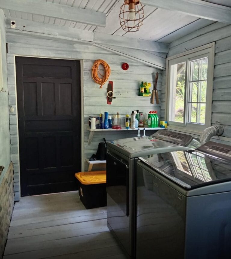 Interior view of a laundry room in a rustic wooden house on Roatan, Bay Islands, Honduras, featuring two washing machines under a window with white-painted wooden walls and hanging tools.