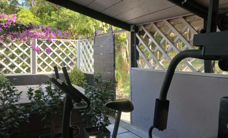 A shaded outdoor patio area on Roatan, Bay Islands, Honduras, with exercise equipment and a view of lush greenery and flowering plants through a white lattice fence.